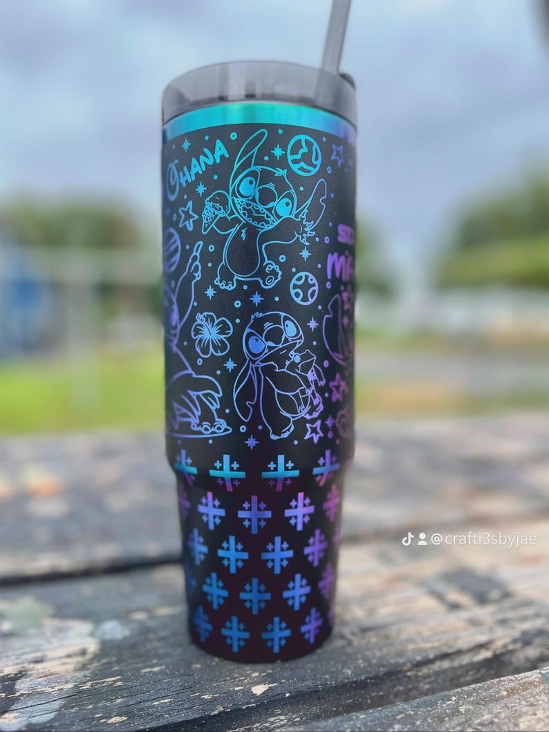 Puede incluir: Vaso negro con tapa y pajita, con un dise&ntilde;o de Stitch de Lilo & Stitch. El dise&ntilde;o incluye la palabra "Ohana" y varias estrellas y formas en azul y morado. La parte inferior tiene un patr&oacute;n repetido de cruces azules y moradas.