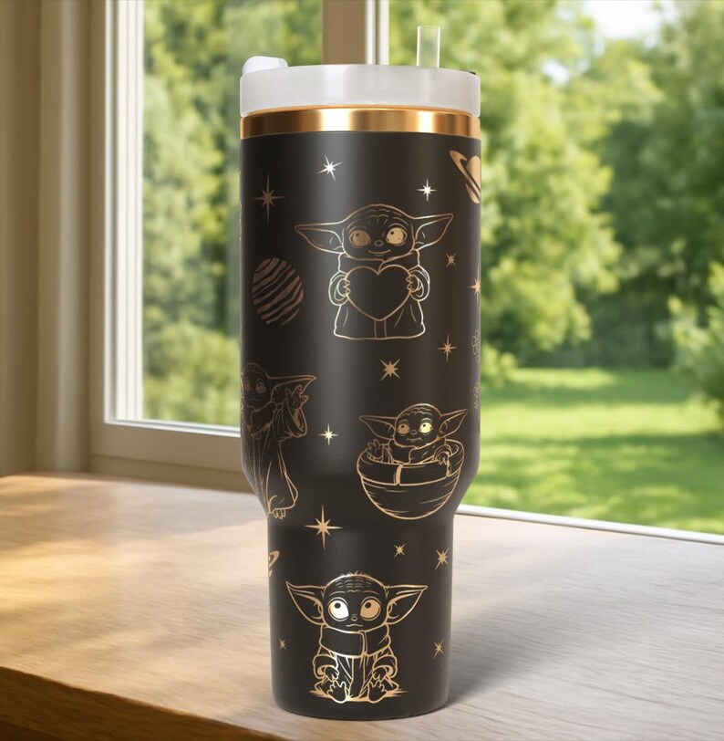 May include: Black tumbler with gold accents and a white lid. The tumbler features gold illustrations of a character holding a heart, in a pod, and other designs. The tumbler has a straw and is sitting on a wooden surface.