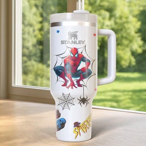 May include: White Stanley tumbler featuring a Spider-Man design. The tumbler has a large handle and a lid with a straw. The design includes Spider-Man, spiderwebs, and the word "Spider-Man". The tumbler is on a wooden surface.