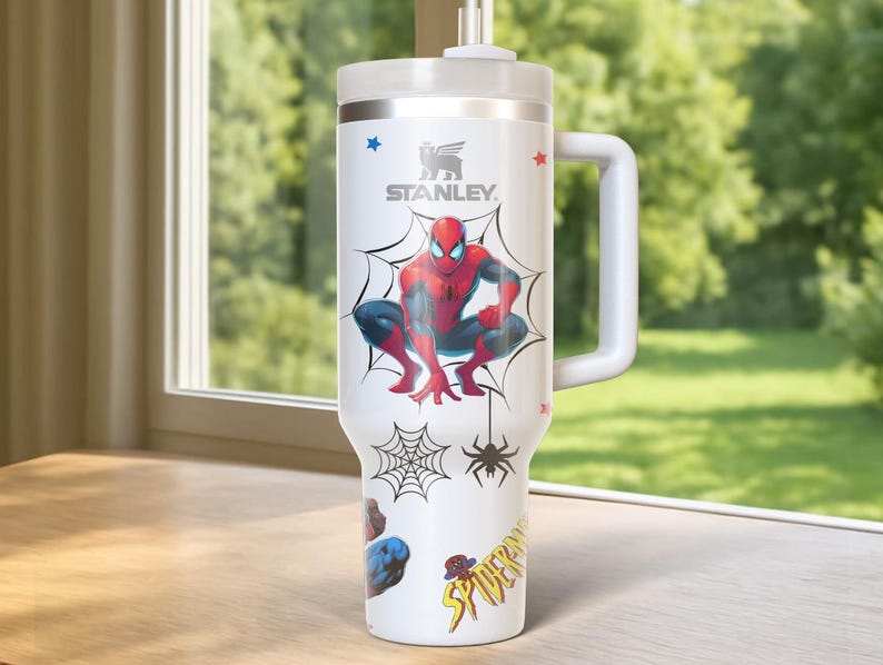 May include: White Stanley tumbler featuring a Spider-Man design. The tumbler has a large handle and a lid with a straw. The design includes Spider-Man, spiderwebs, and the word "Spider-Man". The tumbler is on a wooden surface.