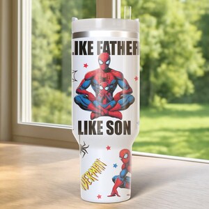 May include: A white tumbler featuring the text "LIKE FATHER LIKE SON" with a graphic of Spider-Man and a child. The tumbler also has a small Spider-Man graphic and a "SPIDERMAN" logo. The tumbler is on a wooden surface.