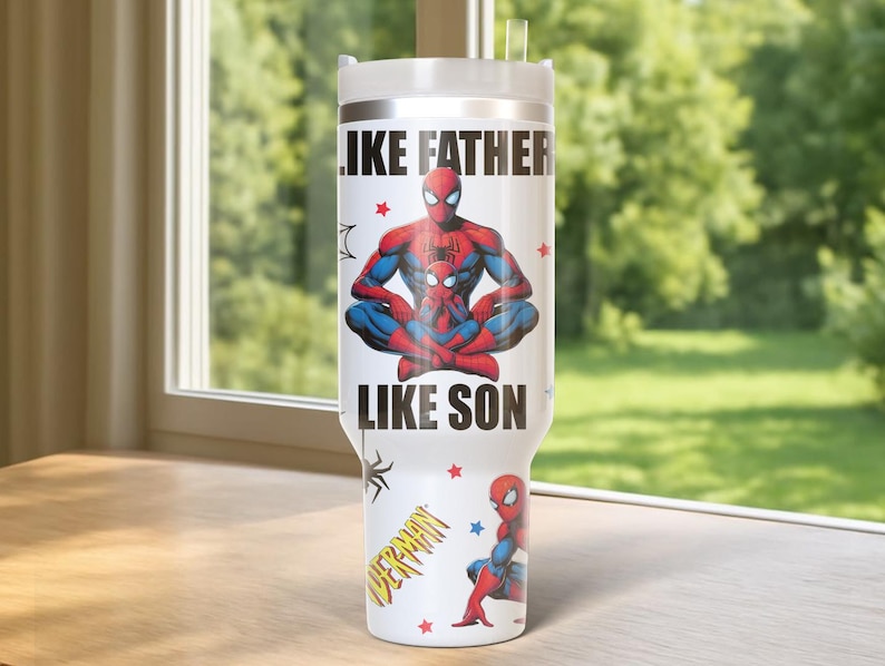 May include: A white tumbler featuring the text "LIKE FATHER LIKE SON" with a graphic of Spider-Man and a child. The tumbler also has a small Spider-Man graphic and a "SPIDERMAN" logo. The tumbler is on a wooden surface.