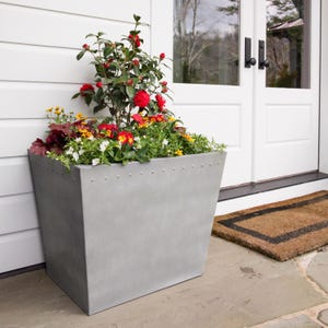 May include: A large, gray metal planter filled with colorful flowers and greenery. The planter has a riveted design along the top edge and is placed on a stone patio next to a white door. The flowers include red, yellow, and white blooms.