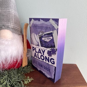 May include: A paperback book titled Play Along by Liz Tomforde from the Windy City Series. The cover features a purple gradient, a baseball, a Las Vegas sign graphic, and poker chips. The page edges are dyed a matching purple color.