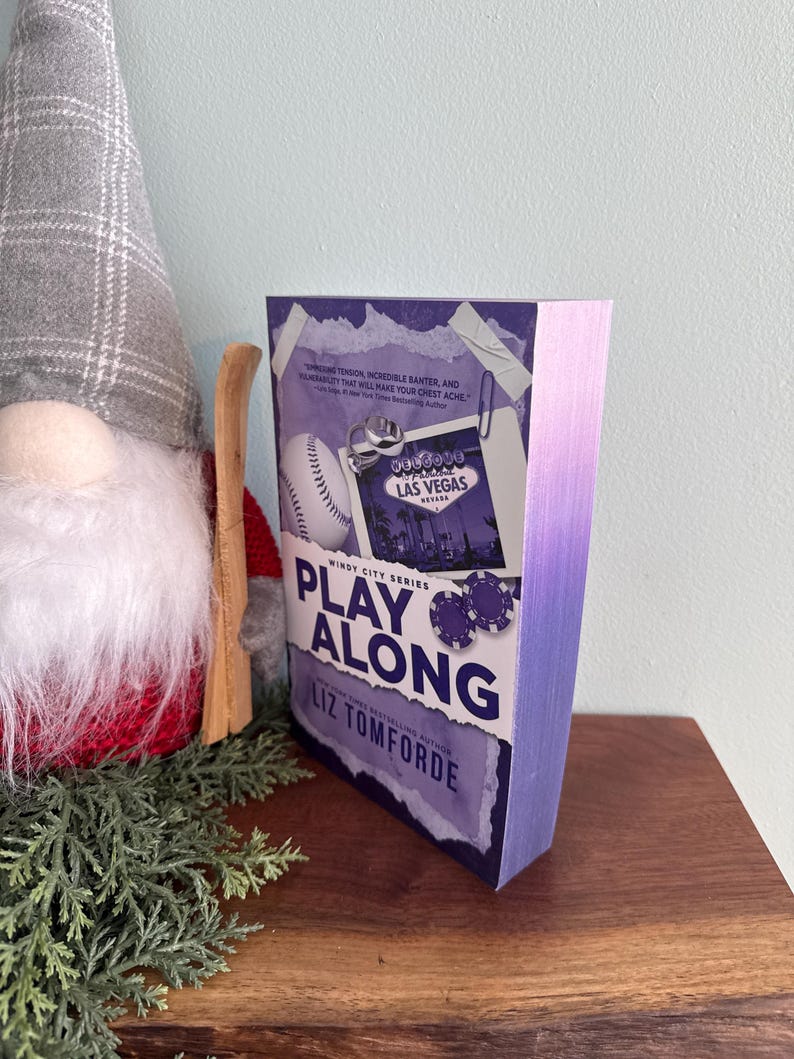 May include: A paperback book titled Play Along by Liz Tomforde from the Windy City Series. The cover features a purple gradient, a baseball, a Las Vegas sign graphic, and poker chips. The page edges are dyed a matching purple color.