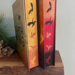 Puede incluir: Dos libros de tapa dura con cubiertas doradas y negras. Los libros tienen un dise&ntilde;o de drag&oacute;n en el lomo. El libro de la izquierda tiene el t&iacute;tulo "The Fourth Wing" en la portada. El libro de la derecha tiene una portada con degradado rojo y naranja.