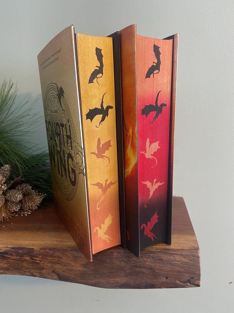Puede incluir: Dos libros de tapa dura con cubiertas doradas y negras. Los libros tienen un dise&ntilde;o de drag&oacute;n en el lomo. El libro de la izquierda tiene el t&iacute;tulo "The Fourth Wing" en la portada. El libro de la derecha tiene una portada con degradado rojo y naranja.