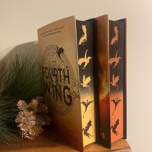 Op de afbeelding: Twee fantasyromans met gouden en oranje omslagen. De omslagen bevatten een draken-silhouetontwerp en de tekst "The Fourth Wing" en "The Fifth Wing".