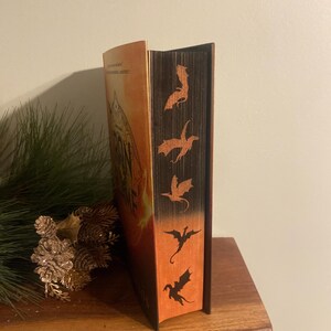 Op de afbeelding: Een hardcover boek met een bruine rug en een oranje omslag. De rug heeft een reeks zwarte draken-silhouetten.