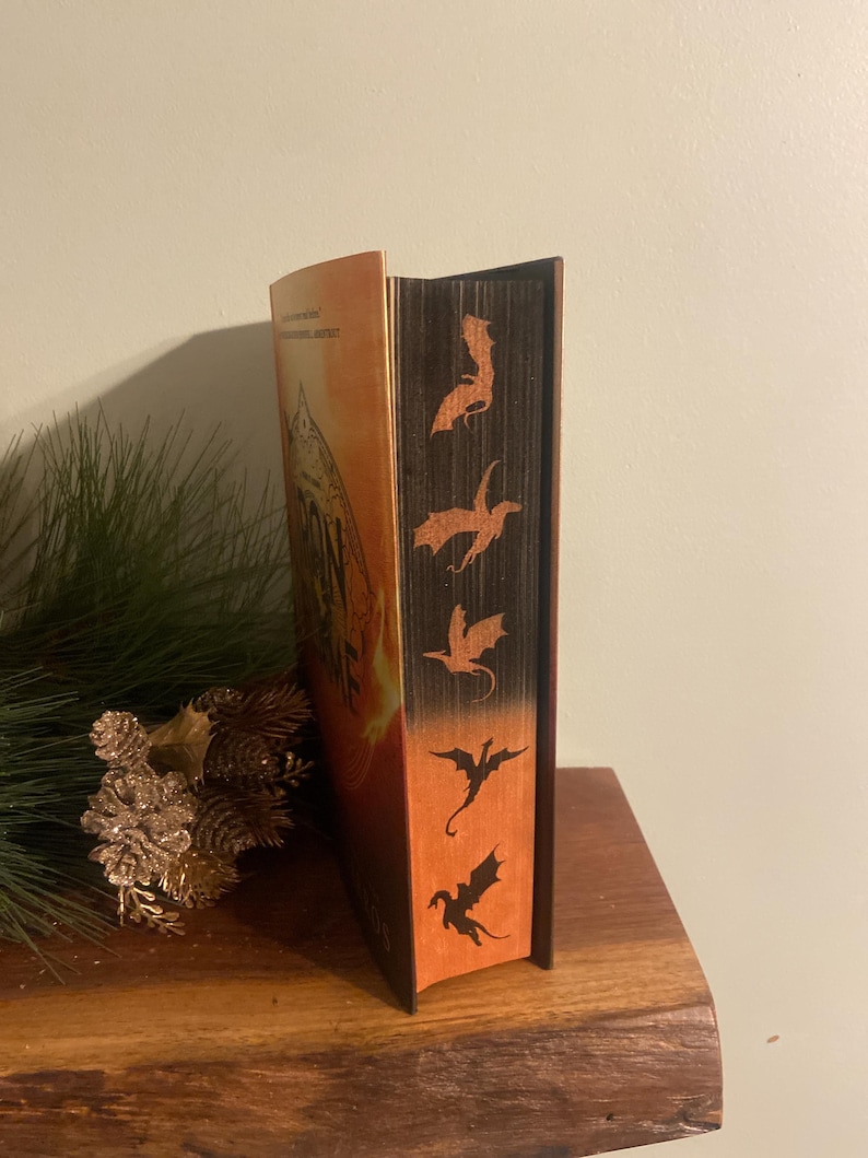 Op de afbeelding: Een hardcover boek met een bruine rug en een oranje omslag. De rug heeft een reeks zwarte draken-silhouetten.