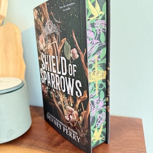 Op de afbeelding: Een hardcover boek getiteld "SHIELD OF SPARROWS" van Devney Perry. De cover heeft een donkere, gedetailleerde illustratie met een kroon, takken en bloemen. De rug heeft een groen bloemenpatroon met gele vogels en roze bloemen.