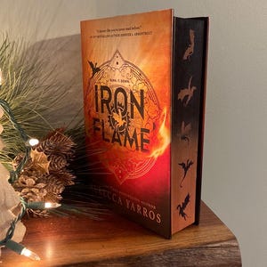 Puede incluir: Un libro titulado "Iron Flame" de Rebecca Yarros, una autora superventas. La portada presenta un fondo naranja y rojo fuego con una silueta de drag&oacute;n y las palabras "Burn it down" en texto negro. El lomo del libro tiene un fondo negro con cinco siluetas de dragones.