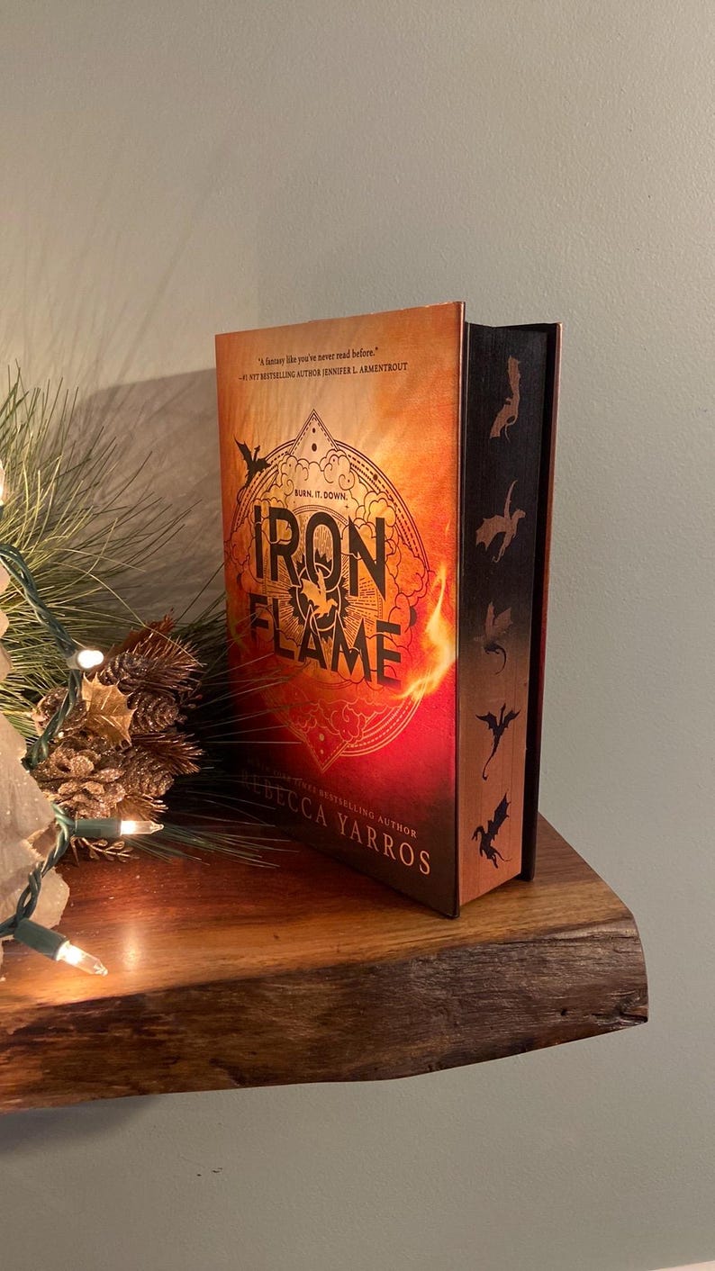 Puede incluir: Un libro titulado "Iron Flame" de Rebecca Yarros, una autora superventas. La portada presenta un fondo naranja y rojo fuego con una silueta de drag&oacute;n y las palabras "Burn it down" en texto negro. El lomo del libro tiene un fondo negro con cinco siluetas de dragones.