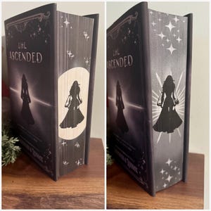 May include: A hardcover book titled "The Ascended" with a dark cover featuring a silhouette of a person. The spine and edges have star and silhouette designs. The book is displayed on a wooden surface.