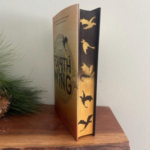 Puede incluir: Un libro de tapa dura en oro y negro titulado "The Fourth Wing" de Rebecca Yarros. El libro tiene un dise&ntilde;o de drag&oacute;n en la portada y una columna vertebral negra con siluetas de dragones dorados.