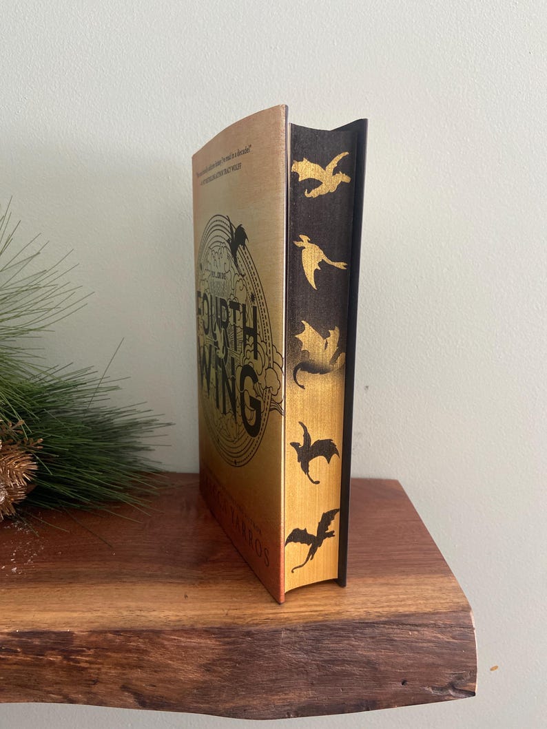 Puede incluir: Un libro de tapa dura en oro y negro titulado "The Fourth Wing" de Rebecca Yarros. El libro tiene un dise&ntilde;o de drag&oacute;n en la portada y una columna vertebral negra con siluetas de dragones dorados.