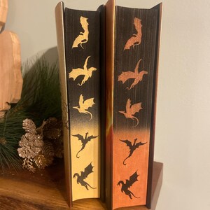 Op de afbeelding: Twee hardcover boeken met draken silhouetten op de ruggen. Het boek links heeft een gouden achtergrond met zwarte draken. Het boek rechts heeft een oranje achtergrond met zwarte draken.