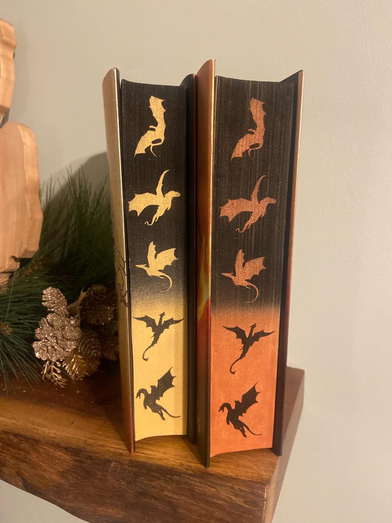 Op de afbeelding: Twee hardcover boeken met draken silhouetten op de ruggen. Het boek links heeft een gouden achtergrond met zwarte draken. Het boek rechts heeft een oranje achtergrond met zwarte draken.
