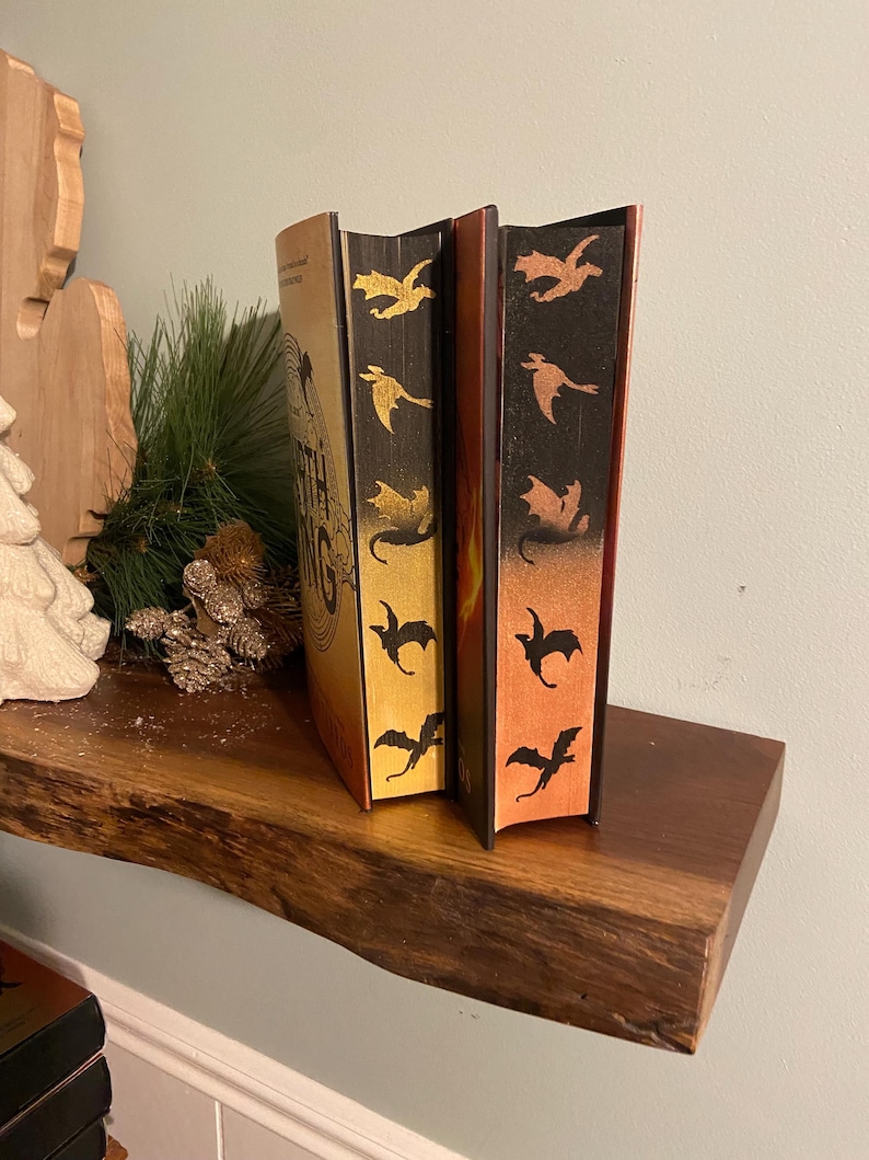 Puede incluir: Dos libros de tapa dura con siluetas de dragones doradas y naranjas en los lomos. El libro de la izquierda tiene el texto "The Fifth" en el lomo. Los libros est&aacute;n en una estanter&iacute;a de madera.