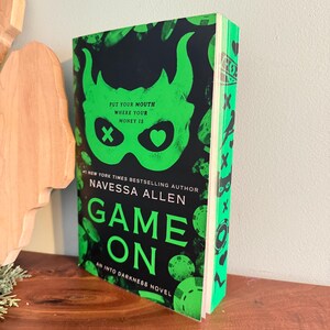 Może przedstawiać: Książka w miękkiej oprawie pod tytułem Game On autorstwa Navessy Allen, z czarną okładką przedstawiającą jaskrawozieloną maskę, żetony do pokera oraz napis Put Your Mouth Where Your Money Is. Grzbiet i krawędzie książki są ozdobione zielonymi symbolami.