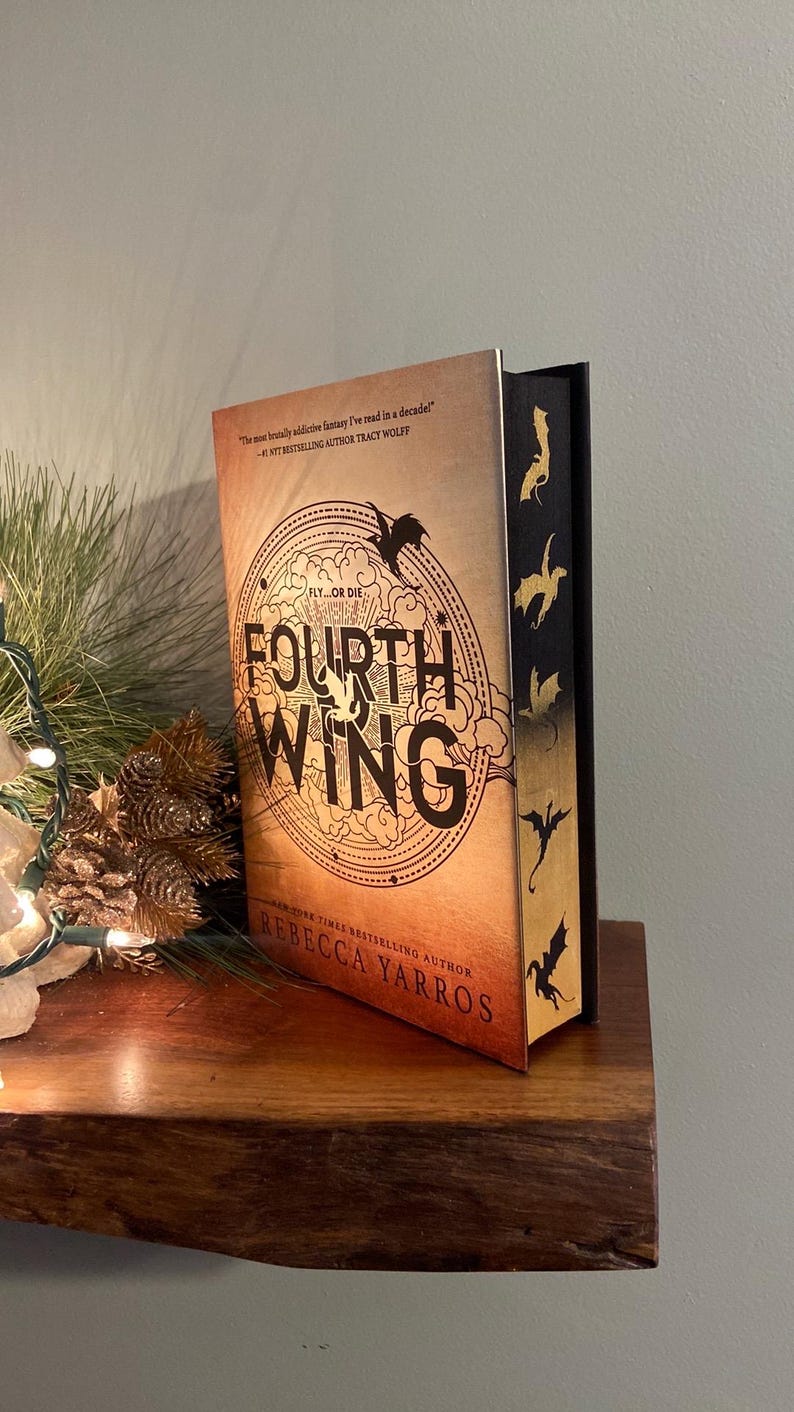 Puede incluir: Un libro titulado "Fourth Wing" de Rebecca Yarros. La portada presenta una silueta de drag&oacute;n y las palabras "Fly or Die". El lomo es negro con siluetas de dragones doradas.