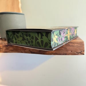K&ouml;nnte beinhalten: Eine rechteckige Box mit einem dekorativen Blumendesign. Die Box hat ein gr&uuml;nes und schwarzes Blumenmuster an den Seiten und ein farbenfrohes Blumendesign auf der Oberseite. Die Box liegt auf einem Holzregal.