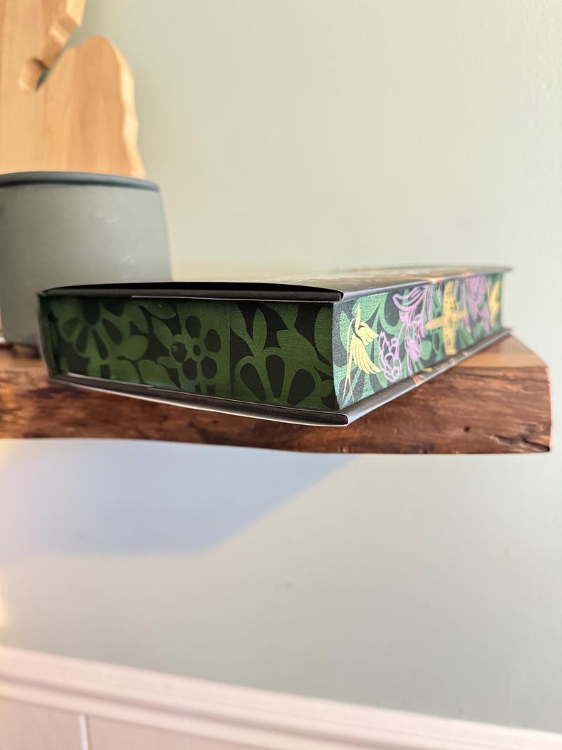 K&ouml;nnte beinhalten: Eine rechteckige Box mit einem dekorativen Blumendesign. Die Box hat ein gr&uuml;nes und schwarzes Blumenmuster an den Seiten und ein farbenfrohes Blumendesign auf der Oberseite. Die Box liegt auf einem Holzregal.
