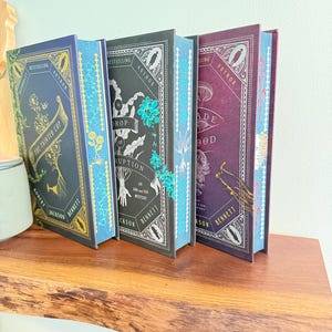 May include: Three hardcover books by Robert Jackson Bennett standing upright. The first is blue titled The Tainted Cup, the second is black titled A Drop of Corruption, and the third is purple titled A Blade of Blood, all featuring intricate gold and white illustrations.