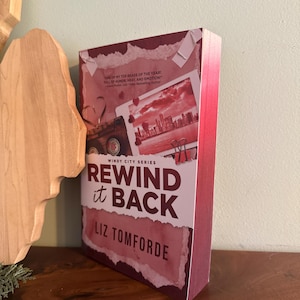 May include: A paperback book titled Rewind it Back by Liz Tomforde from the Windy City Series. The cover features a maroon and white color scheme with a collage of a cassette tape, a city skyline photograph, and red binder clips on a torn paper background.