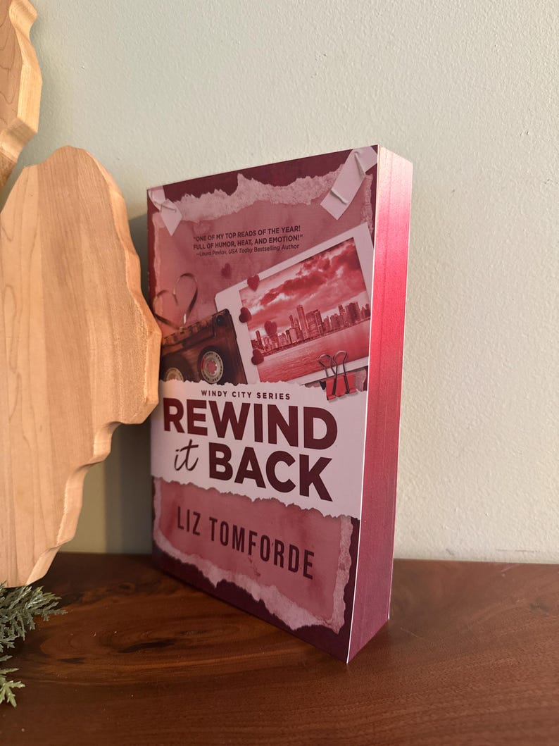 May include: A paperback book titled Rewind it Back by Liz Tomforde from the Windy City Series. The cover features a maroon and white color scheme with a collage of a cassette tape, a city skyline photograph, and red binder clips on a torn paper background.
