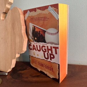 May include: A paperback book titled Caught Up by Liz Tomforde from the Windy City Series. The cover features a baseball, a photograph of a city skyline, and text that reads, Addicting, sexy, and tender in all the right places&mdash;I devoured Caught Up.