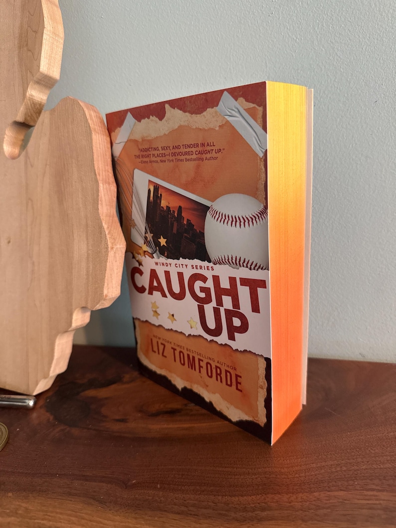 May include: A paperback book titled Caught Up by Liz Tomforde from the Windy City Series. The cover features a baseball, a photograph of a city skyline, and text that reads, Addicting, sexy, and tender in all the right places&mdash;I devoured Caught Up.