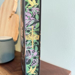 K&ouml;nnte beinhalten: Ein Hardcover-Buch mit einem dekorativen Buchr&uuml;cken mit einem gr&uuml;nen Blumenmuster mit rosa Blumen und gelben V&ouml;geln. Der Titel des Buches ist auf dem Buchr&uuml;cken zu sehen. Das Buch liegt auf einem Holzregal.
