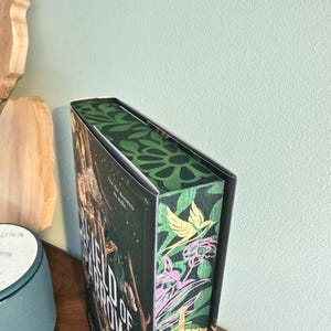 K&ouml;nnte beinhalten: Ein Hardcover-Buch mit dem Titel "Field of Arrows" mit einem dekorativen gr&uuml;nen und floralen Design auf dem Buchr&uuml;cken und den Kanten. Das Cover hat einen dunklen Hintergrund mit goldenen Buchstaben und Bildern. Das Buch liegt auf einer Holzoberfl&auml;che.