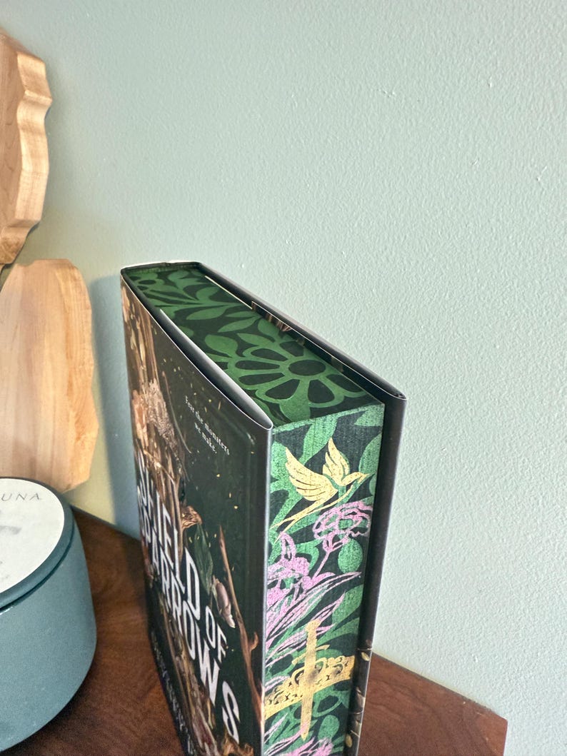 K&ouml;nnte beinhalten: Ein Hardcover-Buch mit dem Titel "Field of Arrows" mit einem dekorativen gr&uuml;nen und floralen Design auf dem Buchr&uuml;cken und den Kanten. Das Cover hat einen dunklen Hintergrund mit goldenen Buchstaben und Bildern. Das Buch liegt auf einer Holzoberfl&auml;che.