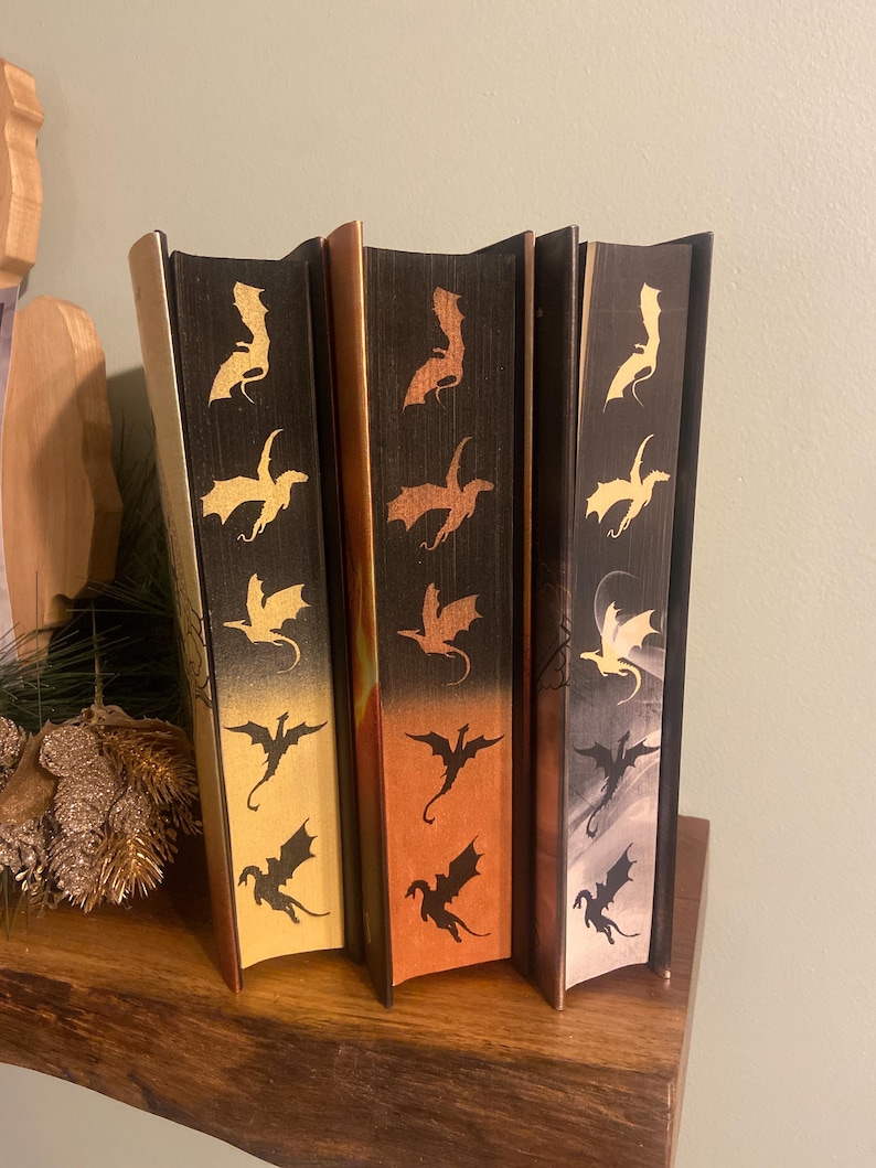 Op de afbeelding: Drie hardcover boeken met zwarte, oranje en grijze omslagen. Elk omslag heeft een herhalend patroon van gouden draken silhouetten.