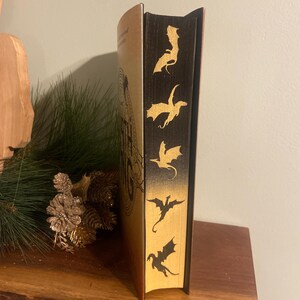 Op de afbeelding: Een gouden en zwart hardback boek met de titel "The Lord of the Rings" op de rug. De rug is versierd met vijf gouden draken-silhouetten.