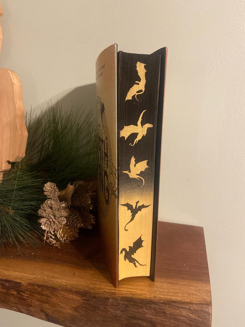 Op de afbeelding: Een gouden en zwart hardback boek met de titel "The Lord of the Rings" op de rug. De rug is versierd met vijf gouden draken-silhouetten.