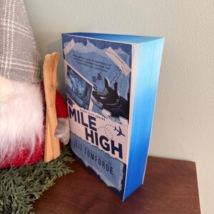 May include: A paperback book titled Mile High by Liz Tomforde from the Windy City Series. The cover features a blue gradient, a graphic of ice skates, a small airplane icon, and text that reads When tension leads to temptation at 30,000 feet.