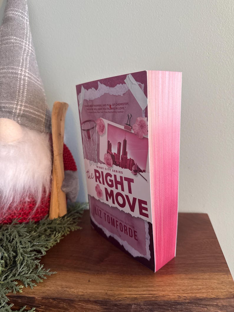 May include: A paperback book titled The Right Move by Liz Tomforde with a pink-tinted cover featuring a city skyline graphic, a basketball hoop, and pink floral accents. The book has bright pink sprayed edges on the pages and a white spine.