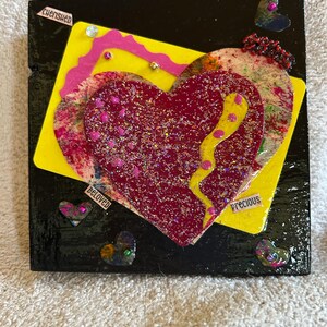 May include: A black wooden square with a red, yellow, and pink heart design. The heart is decorated with glitter and rhinestones. The words "cherished", "beloved", and "precious" are written on the square.