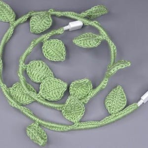 Può includere: Una ghirlanda di foglie all'uncinetto verde avvolta attorno a un cavo di ricarica bianco. La ghirlanda è composta da foglie singole all'uncinetto collegate da un cordoncino sottile.