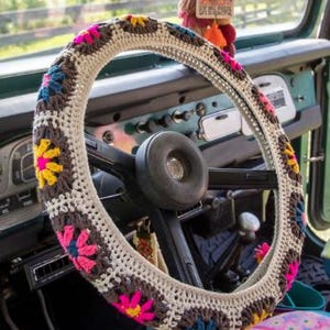 May include: A crocheted steering wheel cover with a floral design in a vintage vehicle. The cover has a cream base with brown, pink, yellow, and blue flower patterns. The steering wheel is black. The cover adds a touch of colour.