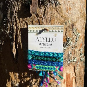 May include: Three colorful friendship bracelets with blue, green, pink, and white threads. The bracelets are displayed on a card with the text "Alyllu Artisans Made in Ecuador".