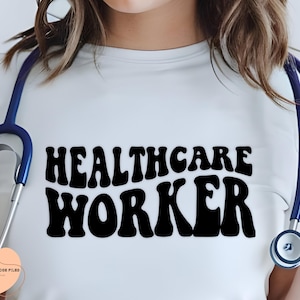 Op de afbeelding: Een wit T-shirt met de tekst "Healthcare Worker" in zwarte, retro-stijl letters. Een blauwe stethoscoop ligt over het shirt.