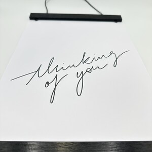 Könnte beinhalten: Schwarz-weiß-Druck mit dem Text "Thinking of you" in kursiver Schrift. Der Druck hängt an einem schwarzen Holzrahmen mit einem Aufhängedraht.