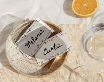 May include: Two white place cards with black cursive text reading "Melanie" and "Curtis" are on a speckled ceramic plate. A half lemon and a glass of water are also in the image. The cards have a decorative black line design.
