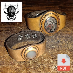 Può includere: Due braccialetti in pelle con bottoni a pressione in metallo e inserti di pietra ovali. Il braccialetto a sinistra ha una pietra bianca, e il braccialetto a destra ha una pietra multicolore.  "Scarab Leatherworks" è stampato su un logo nell'angolo in alto a sinistra.