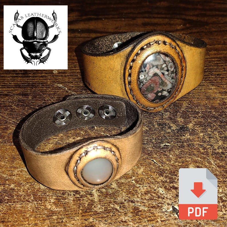 May include: Two leather bracelets with metal snaps and oval-shaped stone insets. The bracelet on the left has a white stone, and the bracelet on the right has a multi-colored stone.  "Scarab Leatherworks" is printed on a logo in the top left corner.