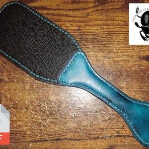 Könnte beinhalten: Ein dunkelbraunes Lederpaddel mit einem blaugrünen Ledergriff. Das Paddel hat einen genähten Rand und ein Loch im Griff. Das Paddel liegt auf einer Holzoberfläche.